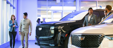 Salesperson and customer walk past Cadillac Escalades in a purple-lit dealership.