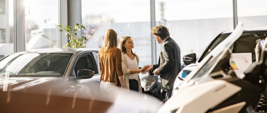Male sales man shaking the hand of a female customer standing next to another female in a car dealership showroom