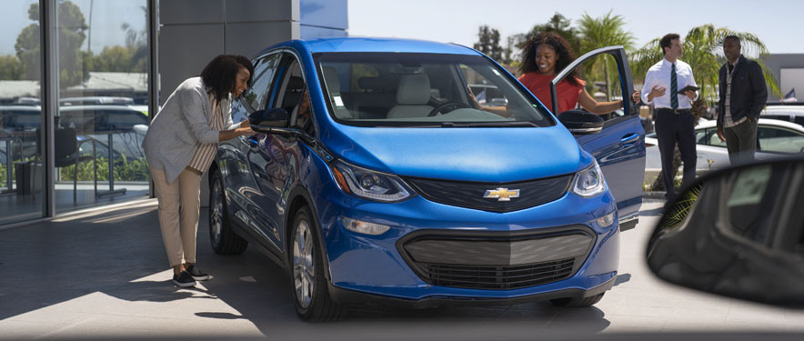  Smiling women checking out blue Chevrolet Bolt at a dealership.