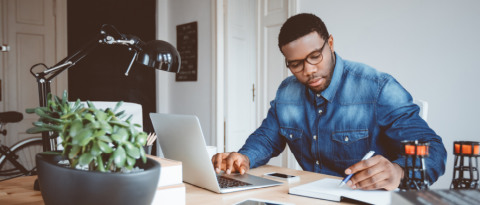 A man at a desk consults a notebook while working on his laptop