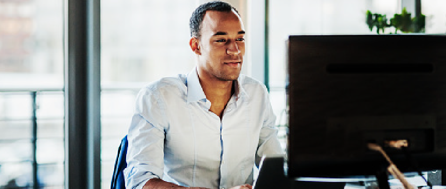 A man smiling while sitting at a desk working on a laptop and looking at a computer monitor 