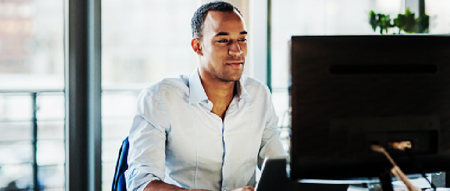 A man smiling while sitting at a desk working on a laptop and looking at a computer monitor