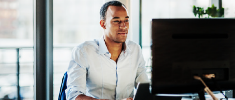 A man smiling while sitting at a desk working on a laptop and looking at a computer monitor