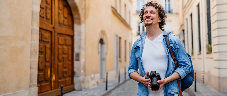 A man with a backpack and camera walks down a European street taking in the sights.