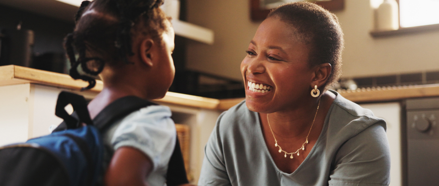 Smiling mother saying goodbye to a young elementary school student with a backpack.