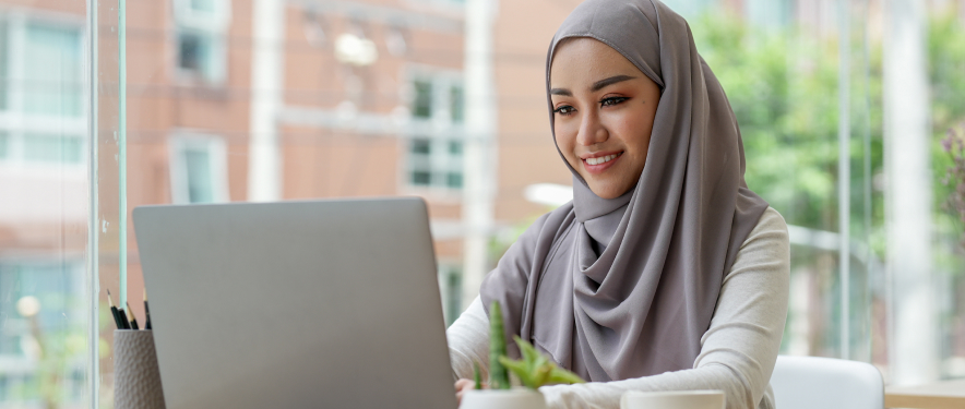 Young adult on a laptop in a glass-walled office building.
